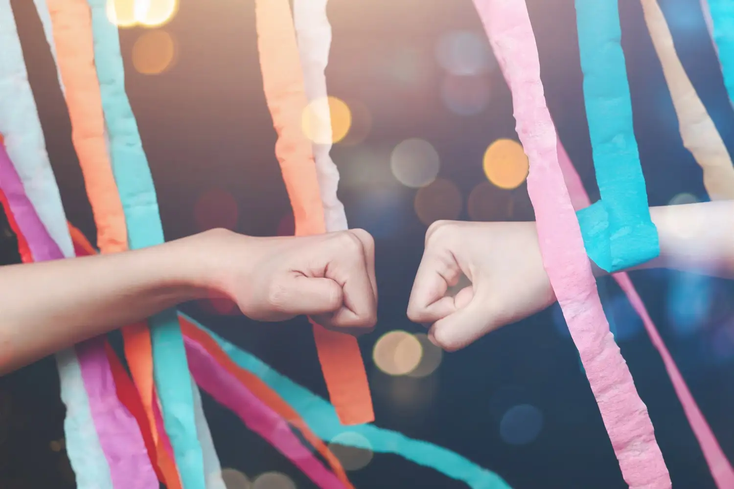 Two hands fist bumping under streamers of a rainbow color